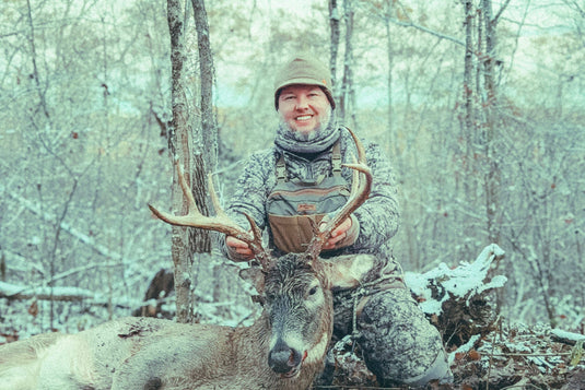 Hunter in ranger green BullPath Moose chest rig kneeling with a whitetail buck in snowy woods.