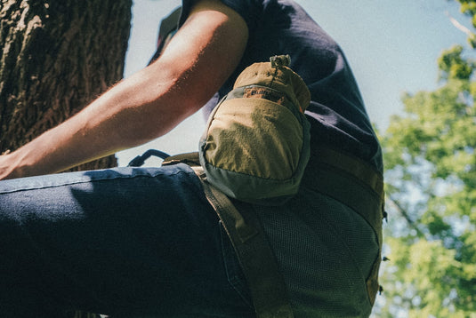 A close-up outdoor shot of a person wearing a BullPath S.A.M X small pouch attached to their El Toro Hunting saddle, with blue jeans and a dark shirt visible, set against a natural background with tree bark and green foliage.